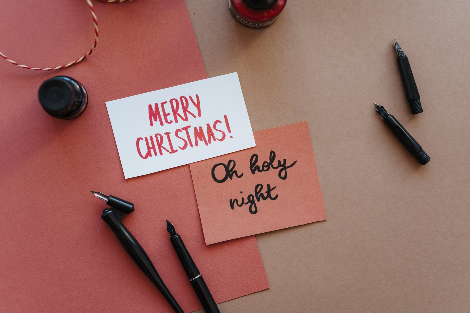 Two hand-lettered cards reading Merry Christmas and Oh holy night lie on red and tan paper beside fountain pens, ink bottles and string.