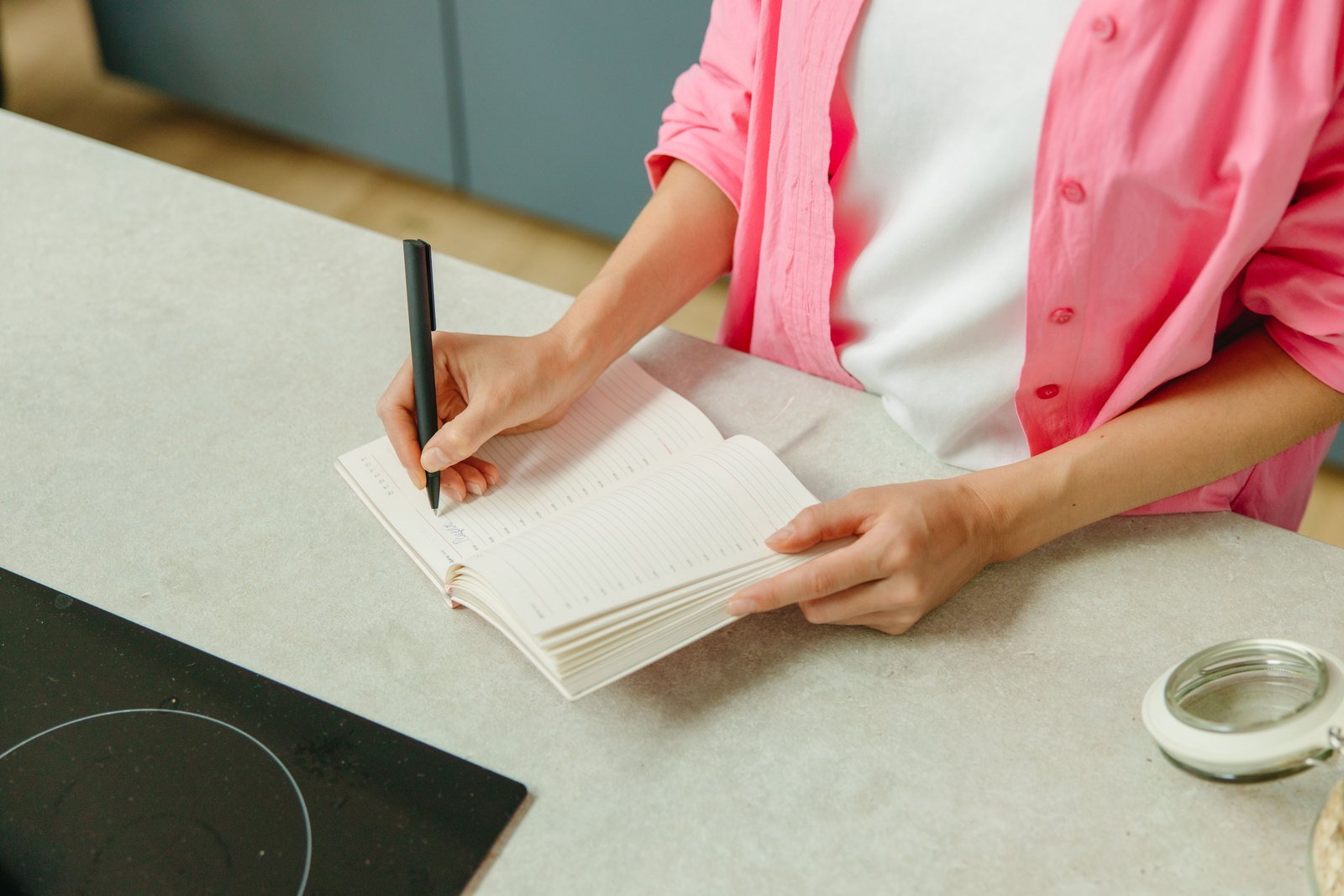 A woman's hands writing a list in an open notebook on a kitchen counter