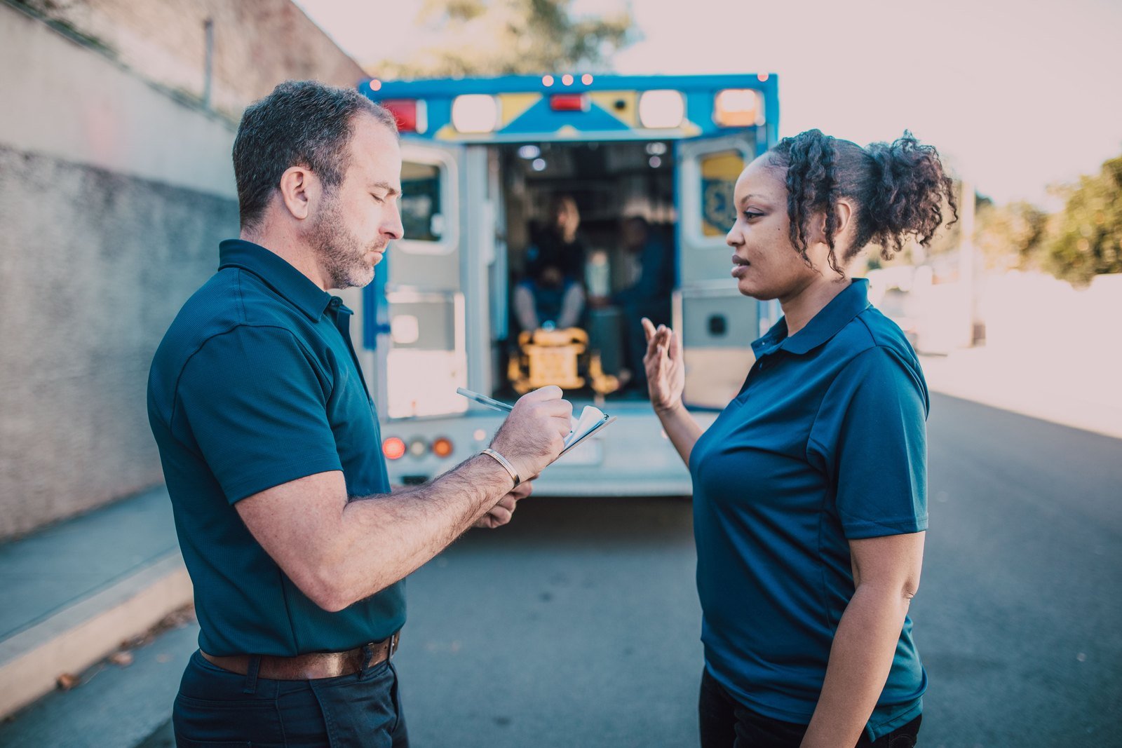 A man writing in a small notepad and a woman talking with one hand raised, standing in front of an ambulance with its back doors open.