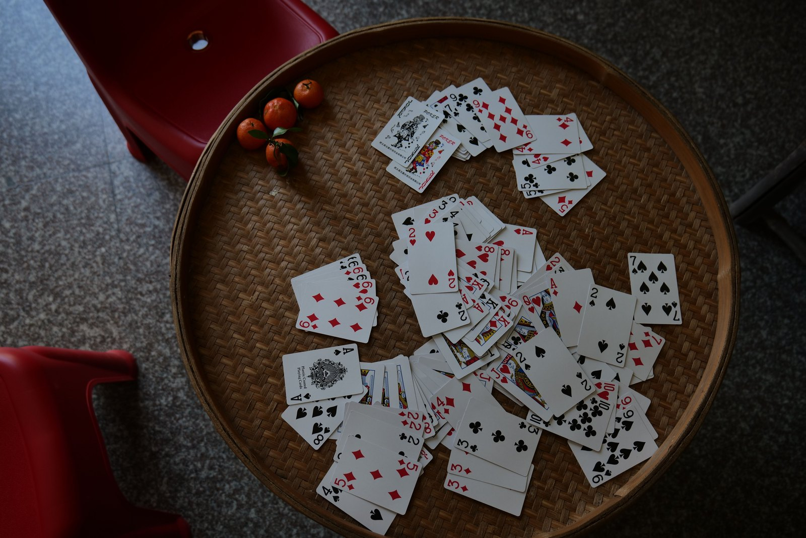 Playing cards scattered face up on a round woven tray table beside four small mandarin oranges, with red plastic stools around it.