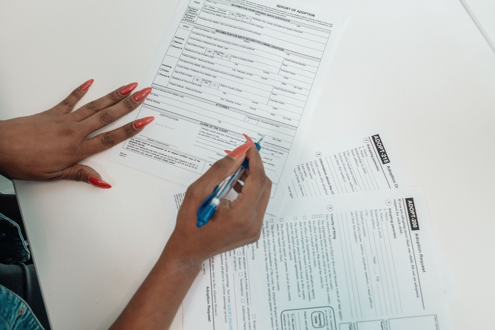 A pair of hands with red painted nails holds a pen over printed forms spread across a white table.