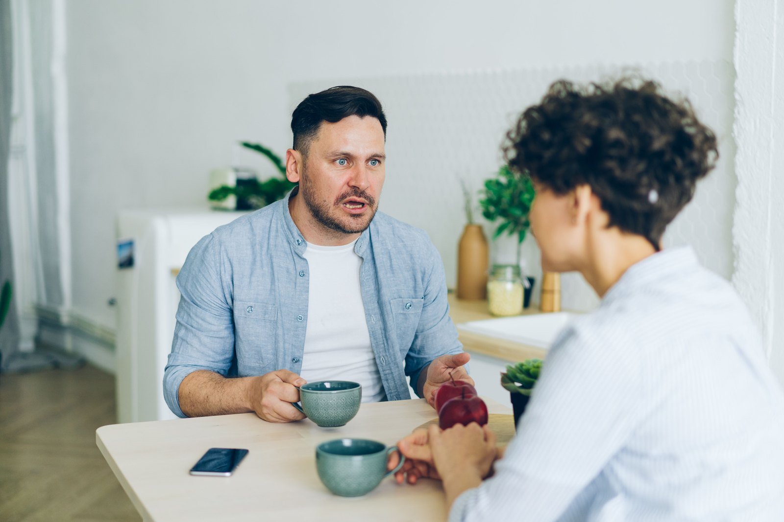 Two people sit across a wooden kitchen table with mugs of coffee between them, mid-conversation