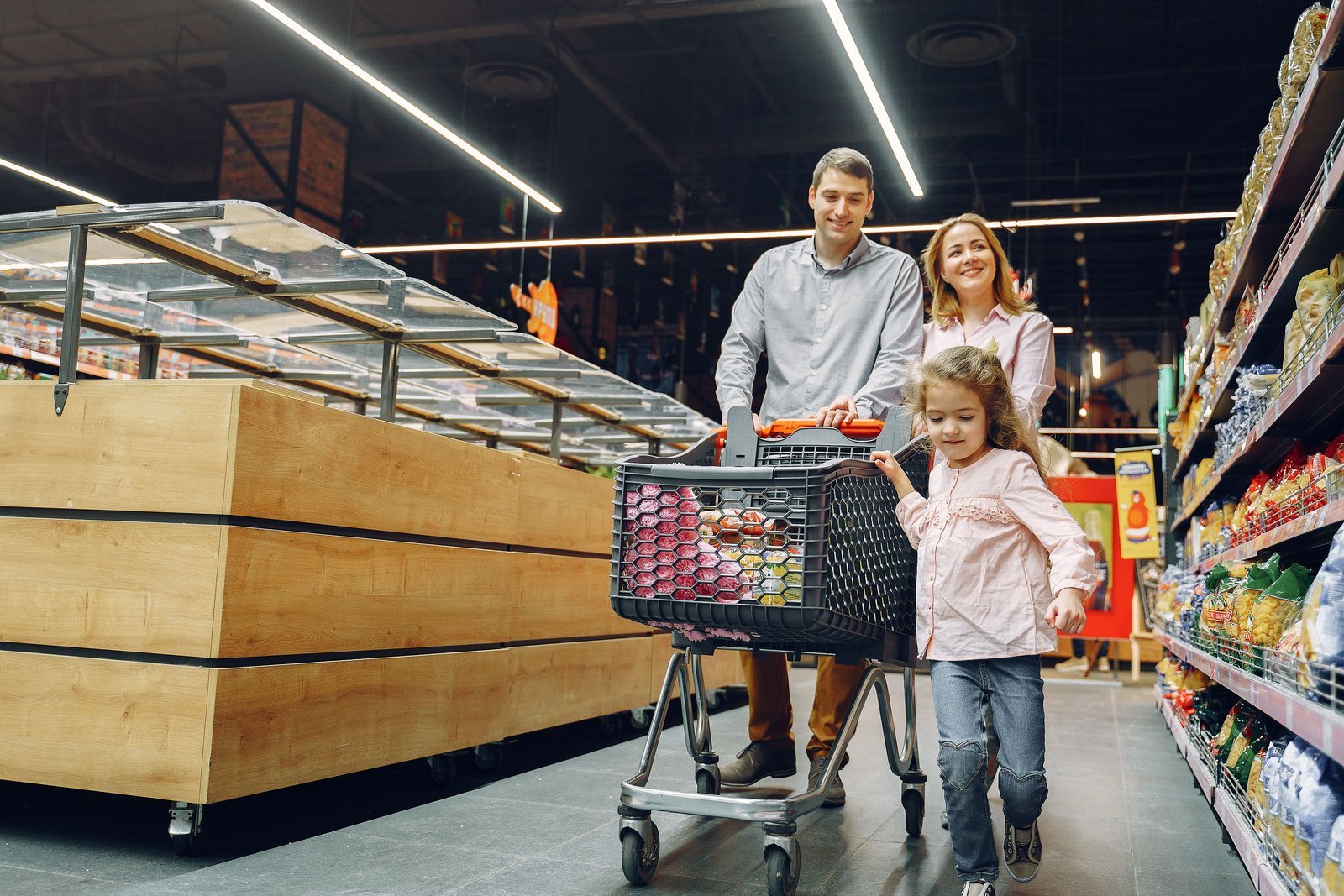 An adult and a child holding hands, walking together toward the exit doors of a busy supermarket, with the checkout lanes and aisles blurred behind them.