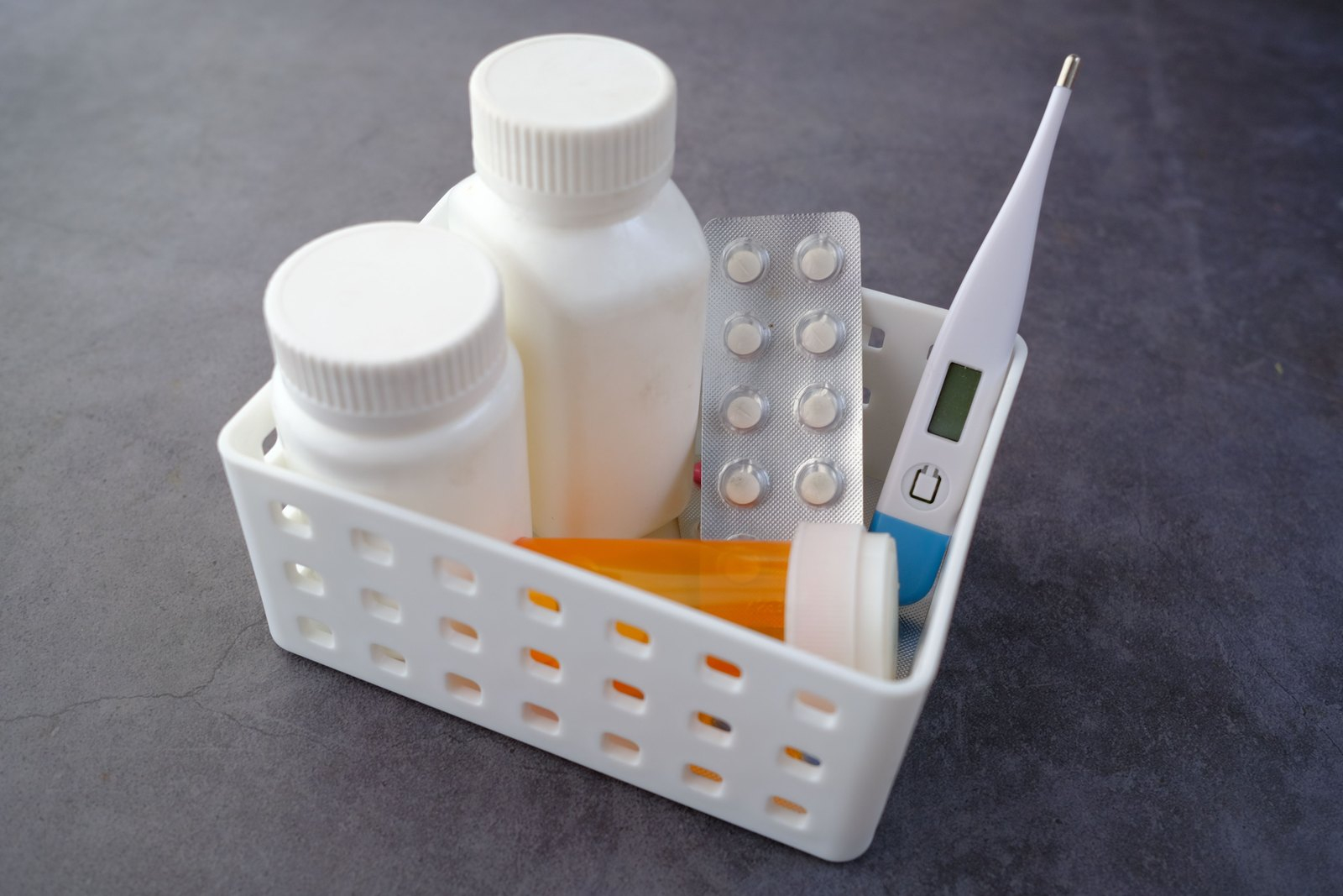 A perforated plastic basket holds two pill bottles, a blister pack of tablets, an orange pill bottle, and a digital thermometer.