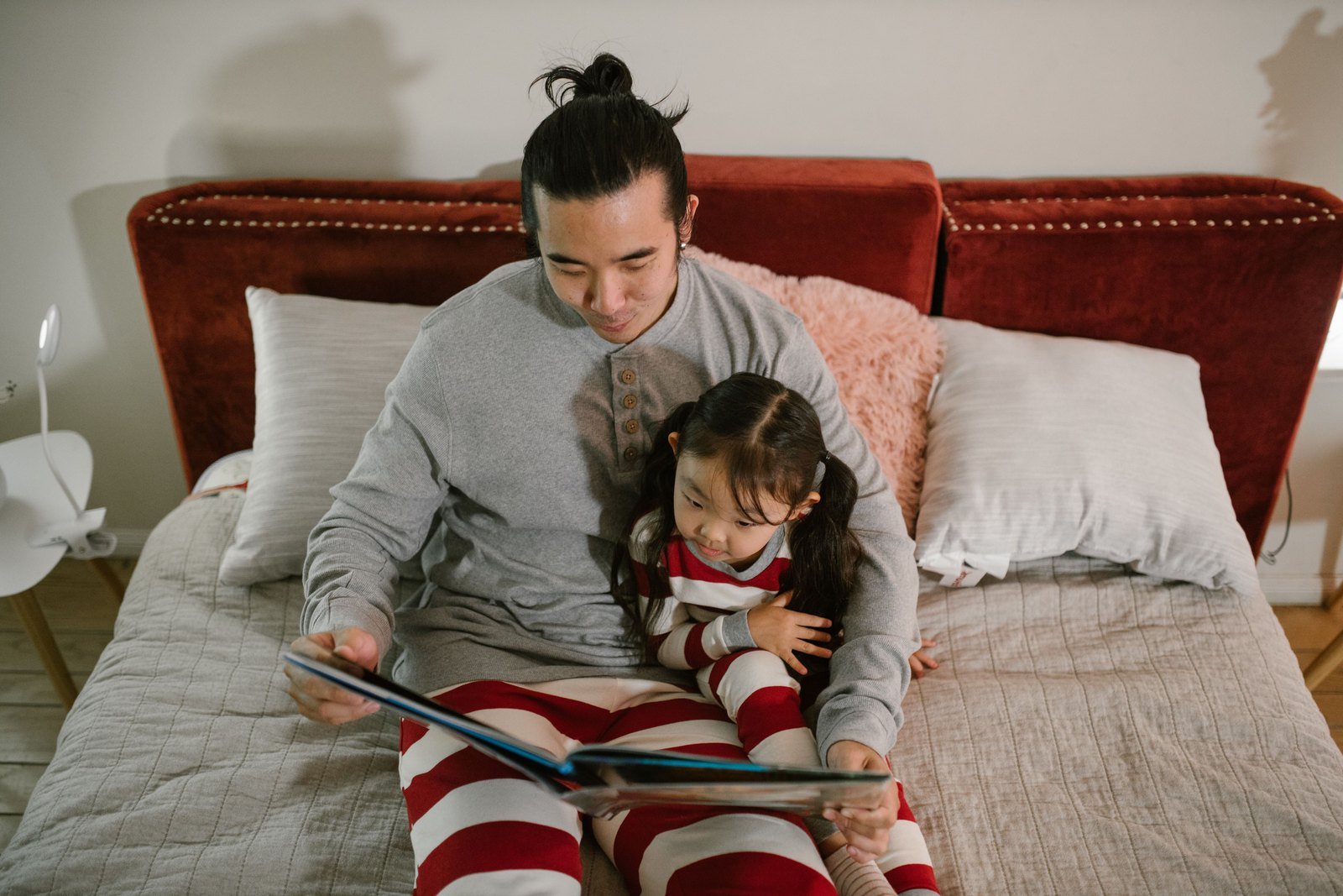 A parent sitting on a bed next to an older child at night, a book open between them under a lamp.