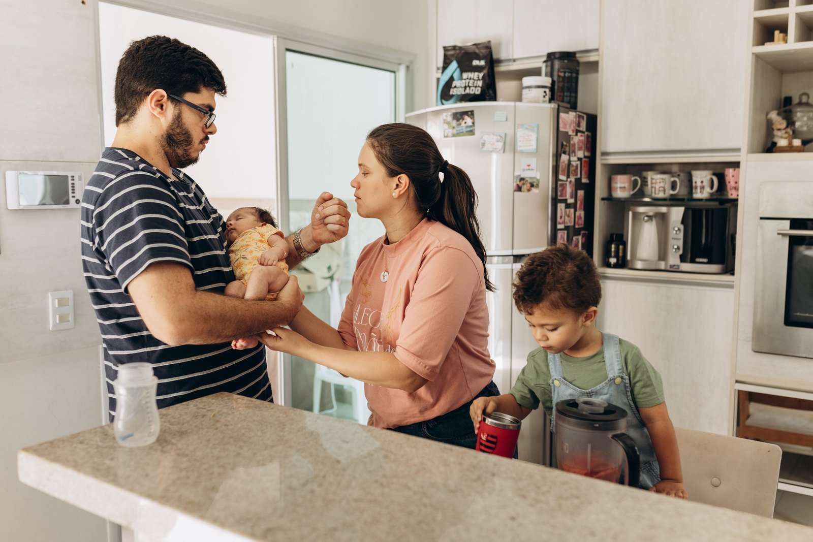 A man holding a baby and a woman stand together in a kitchen as a toddler plays with a small blender on the counter.